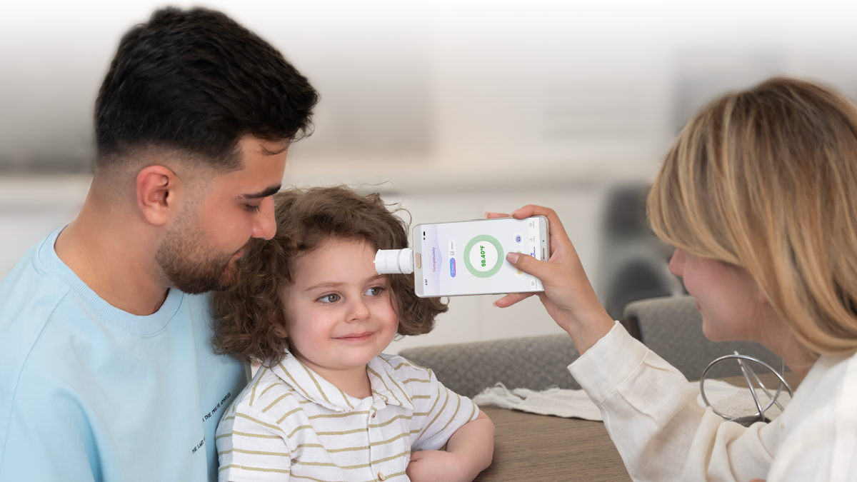 Two parents checking a smiling child’s forehead temperature with TempBuddy plugged into a phone